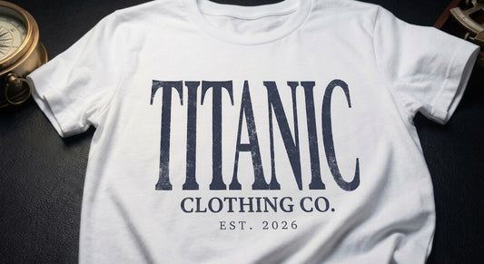 A white short-sleeve T-shirt laid flat on a dark textured surface, featuring large distressed navy text that reads 'TITANIC' with smaller text below reading 'CLOTHING CO.' and 'EST.